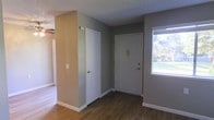 This single-floor residential space offers a straightforward and functional layout designed for comfortable everyday living. The entryway serves as a welcoming transition into the home, featuring rich, dark hardwood-style flooring that extends throughout the main living areas, providing a warm and cohesive aesthetic underfoot. The walls are finished in a calming neutral gray tone, complemented by crisp white baseboards and door trim that add a clean, polished contrast. A textured popcorn ceiling adds a classic touch overhead. Natural light filters into the space through a window near the entry, offering views of the surrounding outdoor greenery and brightening the interior with a pleasant, airy feel. The entryway includes two interior doors, providing access to additional rooms or storage areas, and is thoughtfully designed to manage the flow of the space efficiently. The overall ambiance is clean, understated, and move-in ready, making it a practical and livable environment. The neutral palette and durable flooring choices reflect a space that is both easy to maintain and adaptable to a variety of furnishing and décor styles.