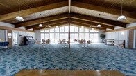 This impressive single-floor gathering hall is defined by its dramatic vaulted ceiling, where rich exposed wood beams and tongue-and-groove paneling rise to a generous peak, creating a warm and airy atmosphere. A sweeping wall of floor-to-ceiling windows spans the far end of the hall, flooding the interior with abundant natural light and offering expansive views of the surrounding wooded landscape. Pendant lights and ceiling fans hang at regular intervals from the beamed ceiling, ensuring both comfort and well-distributed illumination throughout the space. The main hall features a boldly patterned blue floral carpet that anchors the room's central gathering area, while a classic parquet wood floor frames the perimeter, adding a refined, layered quality to the flooring design. Multiple round tables and chairs are arranged across the carpeted area, making the space well-suited for events, meetings, or social gatherings. A piano positioned along one wall adds a cultural and entertainment dimension, while potted plants contribute a touch of natural warmth. The combination of natural wood architecture, generous glazing, and thoughtful furnishing arrangements gives this hall a welcoming, versatile character ideal for a wide range of functions.
