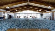 This impressive single-floor gathering hall is defined by its dramatic vaulted ceiling, where rich exposed wood beams and tongue-and-groove paneling rise to a generous peak, creating a warm and airy atmosphere. A sweeping wall of floor-to-ceiling windows spans the far end of the hall, flooding the interior with abundant natural light and offering expansive views of the surrounding wooded landscape. Pendant lights and ceiling fans hang at regular intervals from the beamed ceiling, ensuring both comfort and well-distributed illumination throughout the space. The main hall features a boldly patterned blue floral carpet that anchors the room's central gathering area, while a classic parquet wood floor frames the perimeter, adding a refined, layered quality to the flooring design. Multiple round tables and chairs are arranged across the carpeted area, making the space well-suited for events, meetings, or social gatherings. A piano positioned along one wall adds a cultural and entertainment dimension, while potted plants contribute a touch of natural warmth. The combination of natural wood architecture, generous glazing, and thoughtful furnishing arrangements gives this hall a welcoming, versatile character ideal for a wide range of functions.