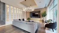 This elegantly designed single-floor amenity space seamlessly blends modern architecture with refined interior finishes throughout its thoughtfully arranged areas. The grand entrance lobby showcases dramatic polished dark marble columns, marble-tiled flooring, and an intricate laser-cut bronze decorative screen, setting a luxurious tone from the moment of arrival. A welcoming seating alcove positioned along floor-to-ceiling glass windows offers sweeping views of a landscaped courtyard and resort-style pool beyond.

The central lounge area serves as the heart of the space, featuring rich wood-plank flooring, a coffered ceiling, and a striking branch-style chandelier that commands attention overhead. An artistic circular-patterned decorative wall panel anchors one side of the room, while a stone-clad media wall with built-in cabinetry provides a sleek entertainment focal point. A generously sized sectional sofa and accent seating create a comfortable and inviting gathering environment.

The fully appointed kitchen island with an integrated cooktop, dual wall ovens, mosaic tile backsplash, and warm wood cabinetry offers a professional-grade culinary experience. Adjacent to this, a formal conference and dining room features a large white table with leather seating, contemporary branch-style pendant lighting, and direct views to the outdoors, making it ideal for private meetings or formal gatherings.