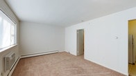This single-floor space presents a clean, straightforward layout centered around a generously sized main room finished with wall-to-wall beige carpeting that lends a neutral, understated feel underfoot. The walls are painted in a crisp white throughout, contributing to a bright and open atmosphere. A baseboard heating unit runs along one wall, providing an efficient and unobtrusive climate control solution. The smooth white ceiling maintains a consistent, uniform appearance across the room. An interior doorway along one wall leads to an adjoining space, offering a glimpse of additional square footage beyond. Electrical outlets are positioned at standard intervals along the walls, and the overall layout is open and uncluttered, providing flexibility for a variety of uses. The simplicity of the finishes and the neutral color palette create a blank-canvas environment that can be adapted to suit different needs and furnishing styles. The space is well-suited for those seeking a functional, low-maintenance interior with a clean aesthetic.