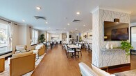 Located at 4121 Concord Creek Place in Richmond, Virginia, this thoughtfully designed single-floor community space offers a generous, open layout that seamlessly blends comfort and functionality. The interior is finished with warm hardwood-style flooring, crown molding, and recessed lighting throughout, creating a polished and welcoming ambiance.

The lounge area anchors one end of the space, featuring stylish sofas, accent chairs, and a marble-top coffee table arranged around a wall-mounted television. Decorative accent walls with botanical-patterned wallpaper and curated artwork add visual interest, while lush indoor plants contribute a lively, organic touch.

The dining area occupies the central and windowed portions of the space, accommodating multiple marble-top tables with upholstered chairs. Large windows dressed with layered curtains flood the room with natural light and offer views of the surrounding community. Decorative columns with coordinating wallpaper accents elegantly define the transition between zones.

The kitchen is well-appointed with white shaker cabinetry, glass-front upper cabinets, grey quartz countertops, a decorative tile backsplash, and stainless steel appliances including a French door refrigerator, dishwasher, and microwave. A direct exterior door provides convenient access to the outdoors, completing this versatile and refined shared space.