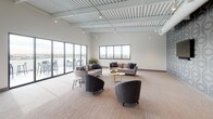 This expansive single-floor communal lounge is designed to accommodate both relaxed socializing and casual gatherings. The open-plan layout is anchored by soaring exposed ceilings with white-painted corrugated decking and industrial-style track lighting, lending a modern, loft-inspired aesthetic throughout the space.

The lounge area is generously appointed with contemporary furnishings, including plush sofas, curved accent chairs in neutral tones, and gold-finished coffee tables arranged to encourage conversation. Large floor-to-ceiling glass sliding doors line one wall, flooding the interior with abundant natural light and providing direct access to an outdoor terrace furnished with bar-height seating and commanding city skyline views.

Toward the opposite end of the space, a distinct dining and bar area features bold geometric patterned wallcovering in deep gray tones, contrasting dramatically with the lighter lounge zone. A sleek bar counter with pendant lighting overhead and amber-toned bar seating creates an inviting setting, complemented by round dining tables and matching chairs nearby. A prominent dark structural column adds architectural definition between the two zones, enhancing the space's visual depth and overall contemporary character.