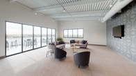 This expansive single-floor communal lounge is designed to accommodate both relaxed socializing and casual gatherings. The open-plan layout is anchored by soaring exposed ceilings with white-painted corrugated decking and industrial-style track lighting, lending a modern, loft-inspired aesthetic throughout the space.

The lounge area is generously appointed with contemporary furnishings, including plush sofas, curved accent chairs in neutral tones, and gold-finished coffee tables arranged to encourage conversation. Large floor-to-ceiling glass sliding doors line one wall, flooding the interior with abundant natural light and providing direct access to an outdoor terrace furnished with bar-height seating and commanding city skyline views.

Toward the opposite end of the space, a distinct dining and bar area features bold geometric patterned wallcovering in deep gray tones, contrasting dramatically with the lighter lounge zone. A sleek bar counter with pendant lighting overhead and amber-toned bar seating creates an inviting setting, complemented by round dining tables and matching chairs nearby. A prominent dark structural column adds architectural definition between the two zones, enhancing the space's visual depth and overall contemporary character.