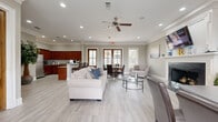 This single-story space is designed as a functional yet welcoming communal or leasing facility, combining several distinct zones within an open, flowing floor plan. Light gray wood-look tile flooring runs throughout, lending a cohesive and clean aesthetic across all areas.

The main lounge area is anchored by a striking white-framed fireplace with a dark stone surround and a wall-mounted flat-screen television above the mantel. Comfortable seating, including a plush white sofa and leather accent chairs, surrounds a round glass coffee table, creating an inviting gathering space. Crown molding and recessed lighting enhance the refined character of the room.

Adjacent to the lounge, a dining area features a round glass-top table with dark leather chairs, positioned near warm wood-framed French doors that allow ample natural light and views of the surrounding exterior. The workstation area nearby includes a modern gray desk and additional seating, suited for administrative tasks.

The full kitchen is well-appointed with rich cherry wood cabinetry, granite countertops, stainless steel appliances, and a peninsula with bar-height seating. A ceiling fan overhead and multiple windows throughout the space reinforce the bright, airy atmosphere. The overall design blends professional functionality with a comfortable, residential warmth.