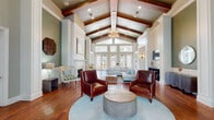 This expansive single-floor gathering space showcases a refined blend of classic and coastal design elements throughout its generously proportioned rooms. The centerpiece of the space is a dramatic great room featuring soaring cathedral ceilings accented by rich, dark wood beams and an elegant sputnik chandelier that anchors the room with a warm, inviting glow. Rich hardwood flooring flows throughout, complementing the sage green and soft white wall palette that lends the space a calm, sophisticated ambiance.

The main lounge area is anchored by a pair of leather armchairs, a light blue sectional sofa, and a round stone coffee table arranged on a powder blue area rug. A classic white-mantled fireplace with a marble surround adds a refined focal point to one wall, while a wall-mounted television on a warm wood panel accent wall is found in an adjacent lounge area. A bank of French doors and transom windows floods the space with natural light and frames views of the surrounding greenery.

A separate dining area features soaring ceilings painted in deep teal, vertical shiplap walls, marble-look tile flooring, and upholstered seating around stone-topped tables. A kitchenette with a stainless steel sink, countertop appliances, and white cabinetry provides practical amenities for gatherings.