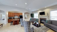 This single-floor space offers an open-concept layout that seamlessly blends comfort and functionality. The living area is thoughtfully furnished with a plush gray sofa accented by teal throw pillows, a pair of bold patterned armchairs, and a dark wood coffee table set atop a textured area rug — creating a warm and inviting gathering space. A wall-mounted flat-screen television and a classic fireplace with a white mantel and stone surround serve as focal points, adding both style and coziness to the room.

Natural light streams in through large windows, brightening the neutral-toned interior and highlighting the vaulted ceiling with recessed lighting that enhances the sense of openness. Decorative arched entryways add an architectural charm to the space, connecting the living area to a well-equipped kitchenette. The kitchenette features warm wood cabinetry, granite-style countertops, a refrigerator, microwave, and a raised breakfast bar with seating — ideal for casual dining or entertaining.

Light-toned tile flooring runs throughout, contributing to the clean and airy feel of the space. An interior windowed vestibule visible in the background hints at a welcoming entryway. Overall, the space balances a residential warmth with practical amenities, making it well-suited for community gatherings or shared use.