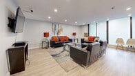 Located at 66 Mooregate Cres in Kitchener, Ontario, this single-floor space offers a thoughtfully designed layout comprising a living room and a fully equipped kitchen. The kitchen features crisp white cabinetry with sleek bar-pull hardware, grey countertops, a white tile backsplash, and stainless steel appliances including a range, microwave, and refrigerator. Light-toned ceramic tile flooring runs throughout the kitchen, and a built-in desk area with matching cabinetry adds a functional touch. Recessed lighting illuminates the space cleanly overhead. The living room is a generously sized, open area finished with light hardwood-style flooring and recessed pot lighting. Large floor-to-ceiling windows along one wall allow natural light to filter in, lending the room an airy, open ambiance. The space is arranged to accommodate multiple seating areas, highlighted by dark leather sofas with vibrant orange accent cushions, a glass-top coffee table, and a wall-mounted television on a geometric console. A white bar counter with bar stools adds a social element near the kitchen transition. The overall aesthetic is modern and clean, with neutral wall tones, contemporary furnishings, and a cohesive design that makes the space feel both functional and inviting.