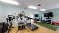 Located at 4013 Wilshire Blvd in Wilmington, NC, this single-story amenity center offers a thoughtfully designed community facility with a bright, coastal-inspired aesthetic throughout. The space is unified by soft aqua-toned walls, herringbone-patterned tile flooring in the common areas, and abundant lighting that creates a clean, welcoming atmosphere.

The entrance lobby greets visitors with a striking sputnik chandelier, French doors that invite natural light, and framed artwork along the walls. A business nook with workstations is conveniently accessible from the entry corridor, separated by frosted glass partitions for a semi-private feel.

A well-appointed reception or kitchenette area features white cabinetry, a dark countertop with an undermount sink, and a large pass-through window overlooking the fitness room, ensuring seamless sightlines throughout the facility.

The fitness area is comprehensively equipped with cardio machines including a treadmill and elliptical, a rack of dumbbells, medicine balls, resistance bands, yoga mats, a weight bench, and a turf training zone. Floor-length mirrors, wall-mounted flat-screen TVs, and decorative artwork contribute to an energizing yet polished environment. Light hardwood-style flooring and access to a restroom complete the fitness space.

A separate office or consultation room features bold teal decorative wall panels, herringbone tile flooring, glass-top tables, and warm accent lighting, offering an elegant and private setting.