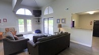 This single-floor space makes a striking impression with its soaring vaulted ceilings and dramatic arched transom windows that flood the interior with natural light. The open living area is the heart of the space, anchored by rich hardwood-style flooring and neutral walls that create a clean, welcoming atmosphere. A wall-mounted flat-screen television is paired with built-in corner shelving, offering a functional yet stylish media setup beneath the arched window feature. The living area is generously sized, comfortably accommodating a large sectional sofa and additional seating, making it ideal for both relaxing and entertaining. Natural light pours in from multiple directions, enhanced by French doors on one side and a wide sliding glass door on the other, both opening to an outdoor deck or patio area. This seamless indoor-outdoor flow adds a sense of expansiveness to the already spacious interior. A large wall mirror further amplifies the open feel of the room. The overall ambiance is airy and casual, with a layout that balances comfort and practicality. The high ceilings, multiple access points to the outdoors, and abundant glazing give this space a light-filled, relaxed character that is both inviting and versatile.