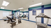 This compact fitness center is thoughtfully designed to maximize its available floor space, providing a dedicated workout environment equipped with essential cardio machines. The room features recumbent bikes, upright stationary bikes, and treadmills arranged to allow comfortable circulation between equipment. A full-length wall mirror runs along one side of the room, visually expanding the space and allowing users to monitor their form during exercise. The flooring consists of speckled terrazzo-style material, offering durability and easy maintenance underfoot. The walls are styled with a bold color scheme, featuring navy blue vertical accent stripes against white walls, topped with a teal horizontal band near the ceiling, creating an energetic and visually distinctive atmosphere. Flush-mounted rectangular fluorescent light panels in the white ceiling provide consistent, even illumination throughout the room. A window along one wall allows natural light to supplement the overhead lighting. Motivational framed signage adorns the walls, reinforcing the space's purpose. A wall-mounted PTAC unit ensures climate control for user comfort. The overall design is clean, functional, and purpose-built to support cardiovascular fitness activity.