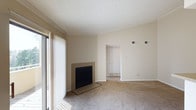 This single-floor residence presents a clean and functional layout with a warm, neutral aesthetic throughout. The living area serves as the heart of the space, highlighted by a fireplace with a sleek black surround set against beige walls, adding a cozy focal point to the room. Large sliding glass doors fitted with vertical blinds allow ample natural light to filter in, brightening the carpeted floors and creating an inviting atmosphere. The textured ceiling adds a classic architectural touch, while crown molding along the upper walls provides a refined finishing detail. Visible HVAC vents are thoughtfully positioned for efficient climate control throughout the space. An open passageway leads from the living area into an adjacent room, suggesting a connected and accessible floor plan that flows naturally between spaces. The soft, wall-to-wall carpeting in a neutral tone runs throughout, contributing to the overall sense of warmth and comfort. The understated color palette of warm beiges and whites creates a versatile backdrop, well-suited for a variety of interior styles. Overall, this space offers a straightforward, comfortable layout with practical features and a welcoming ambiance.