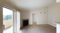 This single-floor residence presents a clean and functional layout with a warm, neutral aesthetic throughout. The living area serves as the heart of the space, highlighted by a fireplace with a sleek black surround set against beige walls, adding a cozy focal point to the room. Large sliding glass doors fitted with vertical blinds allow ample natural light to filter in, brightening the carpeted floors and creating an inviting atmosphere. The textured ceiling adds a classic architectural touch, while crown molding along the upper walls provides a refined finishing detail. Visible HVAC vents are thoughtfully positioned for efficient climate control throughout the space. An open passageway leads from the living area into an adjacent room, suggesting a connected and accessible floor plan that flows naturally between spaces. The soft, wall-to-wall carpeting in a neutral tone runs throughout, contributing to the overall sense of warmth and comfort. The understated color palette of warm beiges and whites creates a versatile backdrop, well-suited for a variety of interior styles. Overall, this space offers a straightforward, comfortable layout with practical features and a welcoming ambiance.