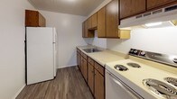 This single-floor unit offers a practical and straightforward layout across several well-defined rooms. The main living area features gray wood-style vinyl flooring and crisp white walls, creating a clean, neutral backdrop. A large window fitted with vertical blinds allows natural light to fill the space, while a wall-mounted air conditioning unit and baseboard heating provide year-round climate control. An adjacent area extends the living space, maintaining the same flooring and bright white walls for a cohesive look.

The bedroom is finished with wall-to-wall carpet in a neutral tone, offering a comfortable and quiet retreat. A window with vertical blinds brings in natural light and an exterior view, while baseboard heating runs along the wall.

The kitchen is designed for efficiency, featuring warm wood-toned cabinetry both above and below the counter, offering ample storage. Light-colored countertops provide workspace alongside a stainless steel sink, a coil-burner electric range, and a full-sized white refrigerator. The same wood-style flooring from the living area continues here, tying the space together.

The bathroom is finished in all white, with small mosaic floor tiles, a combined tub and shower with a curtain rod, a toilet, and a vanity with under-sink storage. A ceiling vent ensures proper ventilation throughout.