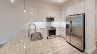 This 1-bedroom, 1-bathroom unit offers a thoughtfully designed single-floor layout that maximizes both comfort and functionality. The open-concept living and kitchen area is anchored by light gray wood-look flooring that flows seamlessly throughout the unit, complemented by crisp white walls that create a bright, airy atmosphere.

The kitchen is a standout feature, showcasing granite countertops, a classic white subway tile backsplash, and sleek white cabinetry extending to the ceiling for ample storage. Stainless steel appliances — including a refrigerator with ice maker, range, and built-in microwave — complete the modern aesthetic, while a peninsula with a single-basin sink and pendant lighting adds both style and prep space.

The bedroom is generously proportioned, filled with natural light from dual windows that offer views of the surrounding neighborhood. A ceiling fan provides year-round comfort. The bathroom features large-format neutral tile surrounding a soaking tub and shower combination, with granite countertops continuing the upscale design language found throughout the unit.

A dedicated laundry area houses a stacked washer and dryer unit alongside a water heater, keeping utilities neatly organized and out of the main living space. The overall ambiance is clean, contemporary, and move-in ready.
