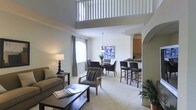 This two-story residence is defined by its dramatic double-height ceiling in the main living area, where an upper-level loft overlooks the space below through a classic white baluster railing. The open-plan layout seamlessly connects the living room, dining area, and kitchen, creating a spacious and cohesive flow throughout the main floor. The living room is anchored by plush, neutral-toned seating arranged around a dark wood coffee table, while large windows dressed with floor-length curtains allow natural light to fill the room. An elegant arched doorway frames the transition into the kitchen, which features a raised breakfast bar with dark bar stools, providing a casual dining option alongside the dedicated dining area. The dining space is centered beneath a pendant light and furnished with a round table and upholstered chairs. Warm beige carpeting runs throughout the main living areas, complementing the neutral wall tones and contributing to a calm, cohesive aesthetic. The upper loft level adds architectural interest and additional functional space to this well-designed, multi-level home.