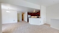 This single-floor residence features an open-concept design that integrates the living area and kitchen into one cohesive space. The living area is finished with neutral wall-to-wall carpeting, creating a warm and comfortable atmosphere throughout. Recessed lighting illuminates the space evenly, complementing the clean white walls and ceilings.

The kitchen is a standout feature, showcasing rich dark wood cabinetry that extends to the upper walls, providing generous storage. A granite-topped peninsula separates the kitchen from the living space, offering both a functional workspace and a natural dividing point between the two areas. Stainless steel appliances, including a refrigerator and microwave, are integrated seamlessly into the cabinetry.

A hallway visible from the main living area leads to additional rooms, with multiple doors suggesting access to private spaces such as bedrooms or a bathroom. The layout is practical and well-organized, with HVAC vents positioned along the lower walls for efficient climate control. Overall, the space presents a clean, functional, and move-in-ready environment suited to everyday living.