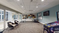 Located at 700 SW 78th Ave in Plantation, FL, this single-floor recreation lounge is a thoughtfully designed communal space built for leisure and social engagement. The room features a neutral-toned carpet that complements the clean white drop-panel ceiling with recessed lighting, creating a bright and welcoming atmosphere throughout.

The space is anchored by a striking feature wall adorned with an eclectic gallery of framed board games, vinyl records, and artwork, lending the room a playful and creative character. A teal accent wall on the opposite side hosts a wall-mounted flat-screen television flanked by additional framed graphic art prints, adding visual depth and a modern touch.

Recreation amenities are central to the layout, including a foosball table, a classic Star Trek pinball machine, and a round card table with seating — offering a variety of gaming options. Comfortable lounge chairs with cushioned seating provide relaxed gathering spots near the floor-to-ceiling glass walls and sliding glass doors, which flood the interior with natural light and provide views of the lush, palm-lined outdoor courtyard beyond. The glass partition walls also connect the lounge visually to adjacent areas of the building, enhancing the open and airy feel of the space.