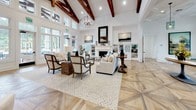Located at 115 Great Lawn Dr in Summerville, SC, this single-story community amenity center offers a thoughtfully designed gathering space with high-end finishes and a cohesive aesthetic throughout. The expansive main clubroom impresses with soaring ceilings adorned with rich exposed wood beams, a geometric chandelier, and abundant natural light streaming through large windows. Comfortable seating arrangements and a relaxed, neutral palette create an inviting atmosphere ideal for socializing.

The catering kitchen is a standout feature, showcasing white shaker cabinetry, quartz countertops, a large central island with bar seating, open wood shelving, and a distinctive geometric-patterned hardwood floor. Pendant lighting and exposed beams lend a warm, farmhouse-inspired character to the space.

A secondary lounge area provides a more intimate setting, anchored by a sleek linear fireplace with floor-to-ceiling tile surround, built-in shelving, and French doors opening to the outdoors. High-top seating near large windows offers views of the surrounding community and palm-lined grounds.

The fitness center is generously equipped with Precor strength machines, treadmills, ellipticals, and cardio equipment, all set on herringbone-patterned flooring. Floor-to-ceiling windows fill the space with natural light and outdoor views, enhancing the workout experience. A dedicated work or lounge nook with dark cabinetry and high-top seating rounds out this well-appointed facility.