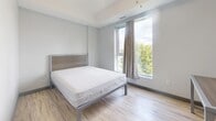 Located at 130 Columbia St W in Waterloo, Ontario, this modern 2-bedroom, 1-bathroom unit offers a well-designed, efficiently laid-out single-floor living space. The open-concept living room is bright and airy, featuring light wood-look flooring, a tray ceiling, recessed lighting, and floor-to-ceiling sliding glass doors that flood the space with natural light and offer elevated views of the surrounding cityscape. The kitchen is thoughtfully equipped with a mix of dark and light cabinetry, granite-style countertops, a white tile backsplash, and full-sized black stainless appliances including a range, dishwasher, and refrigerator. The primary bedroom is generously sized, with large windows framing urban views, light wood flooring, and ample room for furnishings. The second bedroom includes a built-in closet and continues the consistent modern aesthetic with the same light flooring and neutral wall tones. The bathroom features large-format tile flooring, a dark wood vanity with an integrated sink, a wall-mounted mirror with overhead lighting, and clean, minimalist finishes throughout. The unit's overall design is characterized by a cool, neutral palette of greys and whites, creating a contemporary and functional living environment.