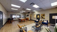 This single-floor amenity facility is thoughtfully designed to offer residents a variety of recreational and social spaces. The centerpiece is a generously sized game and lounge room featuring light wood-look flooring, a drop tile ceiling with recessed panel lighting, and warm neutral walls. A full-size pool table anchors the recreational zone, while a shuffleboard table adds additional entertainment options. Adjacent to the game area, a comfortable lounge zone is furnished with navy blue accent chairs, mustard yellow seating, a patterned area rug, and sleek coffee tables, creating an inviting space for relaxation. Decorative wall art and a mounted flat-screen television enhance the social atmosphere, while large windows dressed with charcoal drapes allow natural light to filter in, offering views to the exterior. A staffed service window and dark-framed doorways connect to administrative areas. The fitness center is equipped with a treadmill, elliptical machine, and additional exercise equipment on rubberized flooring, providing a functional workout environment. Throughout the space, cohesive modern décor, consistent flooring, and well-distributed lighting create a unified and welcoming community amenity area.