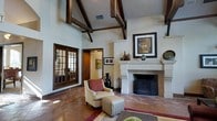 This single-floor space makes a strong architectural statement through its vaulted ceilings adorned with striking exposed dark wood beams, lending a sense of grandeur and rustic elegance to the interior. The main living area serves as the centerpiece of the space, anchored by a substantial stone fireplace with a clean, classic surround that provides both a visual focal point and a warm gathering point. Rich, stained concrete flooring adds depth and character underfoot, complementing the warm tones found throughout the space.

Natural light filters in through a set of French doors with distinctive divided-light panes, framed in dark wood that echoes the ceiling beams and creates a cohesive design language throughout. The doorway leads seamlessly to an adjoining room, reinforcing the open and connected flow of the floor plan.

Decorative touches such as ceramic vessels, framed artwork above the fireplace mantel, and a coordinated seating arrangement with an accent chair and matching ottoman contribute to the space's refined yet approachable aesthetic. The overall ambiance balances traditional architectural elements with comfortable livability, resulting in an interior that feels both distinguished and welcoming.