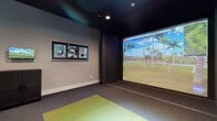 Located at 2600 Executive Dr in Venice, FL, this single-floor indoor golf simulator facility is purpose-built for immersive golf practice and entertainment. The space is divided into two distinct zones that work seamlessly together to deliver a complete simulator experience.

The simulator bay is the centerpiece of the facility, featuring a large-format projection screen that spans the full width of the striking wall, displaying highly detailed, photo-realistic golf course environments powered by an ion3 simulation system. The hitting area is outfitted with a green turf mat set into durable, multi-tone carpet flooring, providing a realistic feel underfoot. The dark ceiling and dark wall paneling surrounding the screen minimize glare and enhance image contrast, while recessed lighting keeps the space well-lit. Golf-themed framed artwork adorns the side wall, adding personality to the room. Entry to the bay is through a set of sleek double barn doors with frosted glass panels, adding a modern architectural detail.

Adjacent to the simulator bay is a comfortable lounge area with natural light filtering through horizontal blinds across multiple windows. Bar-height tables and adjustable swivel stools provide seating for spectators, while a wall-mounted television allows guests to follow the simulation action. A compact storage cabinet and overhead ceiling-mounted projector complete the functional layout of this well-appointed golf entertainment space.
