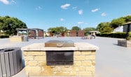 The Game and Barbecue area at Avila 5000 in Sanger is a well-appointed outdoor communal space designed for recreation and social gatherings. At the heart of the space stands a substantial built-in barbecue station, constructed from warm-toned natural stone blocks and topped with a wide concrete surface. A stainless steel grill unit sits atop the structure, providing a functional and durable cooking setup suited for community cookouts and events.

The surrounding courtyard is finished with a smooth concrete surface, offering ample open space for games and activities. A classic wood-and-steel pergola anchors one corner of the area, providing a shaded retreat and a defined gathering spot. Complementing the pergola are additional seating elements, including benches and low tables, distributed throughout the space to accommodate groups of various sizes.

Decorative wood-panel privacy walls add a modern architectural touch and help define the boundaries of the outdoor area. Mature trees and well-maintained hedgerows frame the perimeter, contributing to a sense of enclosure and natural ambiance. The broader community grounds feature grassy lawns, landscaped rock beds, and concrete walkways connecting to the residential buildings. Under open Texas skies, this outdoor amenity space offers a versatile, inviting environment for residents to gather, grill, and enjoy the outdoors.