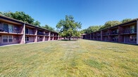 Located at 1435 Nicholasville Rd in Lexington, Kentucky, this residential complex showcases a classic two-story brick exterior arranged around a generously sized, open courtyard. The buildings frame a beautifully maintained central lawn anchored by a large, mature shade tree, creating a welcoming and park-like atmosphere. Paved walkways wind throughout the grounds, connecting units and common areas while bordered by neatly trimmed grass. The complex offers dedicated outdoor amenities designed for communal use, including a built-in gas grill station and a wooden picnic table set on a poured concrete patio — ideal for outdoor gatherings. Individual units feature private balconies accessible from the upper level, providing residents with personal outdoor space overlooking the courtyard and surrounding greenery. On-site parking is conveniently available, with covered carport options also present. The lush tree canopy surrounding the property enhances privacy and shade throughout the warmer months. The overall layout of the complex promotes a sense of community while maintaining a quiet, residential character well-suited to the Lexington neighborhood setting.
