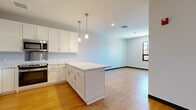 Located at 9 Mill St in Paterson, NJ, this single-floor unit offers a thoughtfully designed open-concept layout with contemporary finishes throughout. The kitchen serves as a central focal point, showcasing white shaker-style cabinetry that extends to the ceiling for generous storage, a large peninsula with marble-look countertops ideal for dining or meal prep, and a full suite of stainless steel appliances including a range, microwave, refrigerator, and dishwasher. Pendant lights hang above the peninsula, adding a polished, modern touch. The kitchen flows seamlessly into an open living area, creating a spacious, connected atmosphere well-suited for both everyday living and entertaining.

Light hardwood flooring runs continuously throughout the unit, contributing warmth and visual cohesion to each space. The additional rooms feature clean, neutral walls with dark-trimmed, grid-style windows that frame outdoor views of the surrounding Paterson streetscape while allowing natural light to fill the interiors. White panel doors and dark baseboards provide crisp architectural contrast. The overall design aesthetic is clean, modern, and move-in ready.