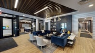 This single-floor space presents a thoughtfully designed communal environment that balances sophisticated aesthetics with functional versatility. The main lounge area serves as a visual centerpiece, anchored by deep navy velvet sofas and soft white accent chairs arranged around a dark coffee table atop a geometric patterned area rug. Light hardwood flooring runs throughout, providing a warm contrast to the darker design elements overhead.

A striking slatted wood ceiling detail defines the lounge zone, complemented by branching brass chandelier fixtures that add an artistic, contemporary focal point. An accent wall clad in dark textured tile houses built-in shelving, lending depth and visual interest to the space. Gold-toned table lamps and marble-top side tables reinforce the refined material palette.

A corridor lined with upholstered booth-style seating pods offers more private, semi-enclosed areas suitable for focused work or small meetings. Glass-panel partitions and black-framed doors maintain visual connectivity while defining separate zones. Near the entrance, floor-to-ceiling windows flood the space with natural light and offer views of the surrounding outdoor landscape. A reception or desk area is visible in the background, completing the functional layout of this well-appointed communal space.