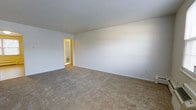 This single-floor residence features two bedrooms and one bathroom within a thoughtfully arranged floor plan. The main living area, pictured here, is a spacious, open room finished with wall-to-wall neutral carpeting that adds warmth underfoot. Crisp white walls and a clean white ceiling create a bright, airy atmosphere, providing a versatile canvas for a variety of furnishing styles. The room offers ample space for a full living arrangement, and a doorway visible in the background connects seamlessly to the adjacent areas of the home, suggesting a practical and flowing layout. The clean lines and understated finishes throughout give the space a move-in-ready appeal. The two bedrooms offer private retreats, while the shared bathroom serves the home efficiently. The overall design is straightforward and functional, emphasizing usable space and neutral tones that allow residents to personalize the interior with ease. The single-floor layout ensures convenient accessibility throughout, making it a practical and comfortable living option.
