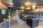 This single-floor amenity space delivers a vibrant, multi-use environment designed for both recreation and relaxation. The interior showcases a bold industrial aesthetic, with dark-painted ceilings, exposed structural elements, textured accent walls with marbled finishes, and warm brick-pattern tile flooring throughout.

The lounge area anchors the space with plush dark leather and upholstered sofas arranged around a low coffee table, accented by striking amber pendant lights that cast a warm glow. A wall-mounted flat-screen TV and high-top bar tables with stools provide additional gathering spots for socializing or casual entertainment.

A full-size ping pong table occupies a central zone, adding a playful recreational element to the space. Nearby, a dedicated bike storage wall features a row of mounted vertical bike racks, offering convenient and organized storage for cyclists.

A roll-up garage-style door connects to an adjacent open flex area with lighter gray walls and recessed lighting, suitable for a variety of activities. Large sliding glass doors in the lounge zone invite natural light and provide views of the surrounding outdoor greenery. The seamless flow between zones makes this a highly functional and energetic communal destination.
