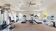 This fitness center is a comprehensively outfitted single-story space designed to support a wide range of workout routines. The room features a tray ceiling with a ceiling fan, contributing to both the sense of height and air circulation throughout the space. Neutral-toned walls and carpeted flooring give the interior a clean, functional aesthetic, while multiple windows along the perimeter flood the room with natural light and offer views of the surrounding landscaping.

Cardio equipment is prominently featured, including two treadmills, an elliptical trainer, and an upright stationary bike, all arranged to allow comfortable spacing between users. A multi-station cable machine and a dedicated free weights area with a tiered dumbbell rack and adjustable bench provide strength training options. Additional functional fitness accessories include medicine balls, stability balls, yoga mats, and a SmartMat core exercise board.

The space is accessed through a set of glass-paned double doors, and wall-mounted mirrors enhance the sense of openness. Security cameras and posted fitness center rules reflect a well-maintained, community-oriented facility. The overall layout is spacious and thoughtfully organized to accommodate multiple users simultaneously.