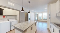 This well-designed single-floor residence offers a thoughtfully laid-out open-concept floor plan that seamlessly connects the kitchen, dining, and living areas. The kitchen serves as the heart of the home, showcasing crisp white shaker-style cabinetry with matte black hardware, polished granite countertops, a subway tile backsplash, and a full suite of stainless steel appliances including a French door refrigerator, electric range, over-the-range microwave, and dishwasher. A generously sized central island with matching granite provides additional prep space and seating, illuminated above by a pair of stylish industrial pendant lights. Light wood-toned flooring flows throughout the main living spaces, complementing the bright, neutral palette of the walls and ceilings fitted with recessed lighting. Large windows in the living area invite abundant natural light and offer views of the surrounding exterior. A dedicated in-unit laundry area is conveniently tucked nearby. The bedroom features plush carpeting, a ceiling fan, and windows overlooking the outdoors. The bathroom is finished with a granite-topped white vanity, wood-look flooring, and direct access to a spacious walk-in closet, completing this clean and functional living space.