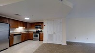 This single-floor space showcases an open-concept layout where the kitchen flows seamlessly into the living area. The kitchen is well-appointed with rich, warm-toned wood cabinetry offering generous storage space, complemented by stainless steel appliances including a freestanding range with microwave above. A sink is positioned along the cabinetry runs, and light-toned tile flooring grounds the kitchen area. The transition into the living space is marked by a shift to neutral carpeting, creating a comfortable and defined zone for relaxation. A set of classic double doors with six-panel detailing and brass hardware adds a traditional architectural touch, likely leading to a closet or storage area. Recessed and flush-mount ceiling lighting provides consistent illumination throughout. The neutral white walls and ceiling contribute to a bright, clean aesthetic, while the overall layout makes efficient use of the available space. The design is practical and cohesive, making it well-suited for everyday living.