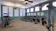 This fitness center offers a spacious, single-floor layout designed to accommodate a range of exercise needs. The room is painted in a calming teal-green hue, complemented by white trim and a neutral carpet-tile floor that provides both comfort and durability underfoot. A row of large windows lines the upper portion of the walls, flooding the space with natural light while maintaining privacy. Multiple ceiling fans are suspended from the elevated ceiling, ensuring consistent air circulation throughout workouts. The cardio section is well-stocked with several treadmills and elliptical machines arranged in an open, accessible formation. A wall-mounted television is positioned for easy viewing during cardio sessions. Full-length mirrors line one wall, visually expanding the space and allowing users to monitor their form. Additional strength and resistance equipment is visible toward the rear of the room. The high ceiling height enhances the sense of openness, contributing to a comfortable and motivating workout environment. Overall, the fitness center combines functional equipment with a clean, organized layout and an airy atmosphere.