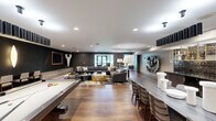 Located at 155 E Broad St in the heart of Columbus, Ohio, this single-floor amenity space offers a refined and contemporary environment designed for both socializing and relaxation. Rich hardwood flooring flows throughout, tying together two distinct yet complementary zones within the space.

The entertainment wing features a full billiards table, a long communal dining or game table with woven chairs, and a dramatic dark-walled media lounge. Deep charcoal walls anchor the lounge area, which is equipped with built-in countertop seating beneath dual flat-screen televisions and a sculptural wall sconce. A large sectional sofa, bold cowhide-patterned area rug, and curated art pieces give this area a luxurious, club-like ambiance. A sleek wet bar with mirrored tile backsplash is visible in the background.

The second zone transitions into a bright, airy lounge characterized by soaring floor-to-ceiling windows framed in dark steel, offering sweeping views of the surrounding Columbus cityscape and lush greenery. Decorative textured ceiling panels and oversized drum pendant lights add architectural character. Intimate seating arrangements with blue-toned sofas, natural wood accent chairs, and carefully selected artwork create an inviting retreat filled with natural light.