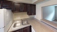 This single-floor unit at River City Place offers a straightforward, functional layout designed for comfortable everyday living. The living area is finished with wall-to-wall carpet in a neutral beige tone, complemented by warm taupe walls, white baseboards, and a textured popcorn ceiling. A wall opening with a pass-through ledge connects the living space to the kitchen, enhancing the sense of openness between the two areas. Wall-mounted sconce lighting adds a warm ambiance to the main living zone, while the entryway transitions to durable hardwood-style flooring that extends through the hallway.

The kitchen is equipped with rich, dark espresso-toned cabinetry featuring sleek bar-pull hardware, paired with speckled laminate countertops that provide ample workspace. A double-basin stainless steel sink sits beneath upper cabinets, offering practical storage throughout. A white refrigerator is visible through the pass-through opening.

The bedroom is generously proportioned, with soft carpet underfoot and a window fitted with horizontal blinds that allow natural light to filter in. The bathroom includes a full tub-shower combination, a window for natural ventilation, wood-style flooring, and a vanity with a countertop. The overall palette throughout the unit is cohesive, with neutral tones creating a calm and adaptable living environment.