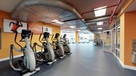 The Fitness Center at 100 Commons Park North in Stamford, Connecticut, is a fully equipped, single-floor facility designed to support a comprehensive workout experience. The open layout maximizes the available floor space, providing ample room for both cardio and strength training activities without feeling crowded.

The cardio area features a row of elliptical machines positioned along one wall, accompanied by treadmills visible toward the rear of the space. Strength training equipment, including a cable and pulley machine, is situated toward the far end of the room, offering a variety of resistance-based exercise options. The flooring throughout is a durable rubber material, ideal for high-traffic fitness environments.

Large windows along the far wall invite abundant natural light, creating an energizing atmosphere while offering views of the surrounding area. The industrial-style ceiling features exposed ductwork and bright red piping, lending the space a dynamic, modern aesthetic. Recessed and flush-mounted lighting supplements the natural light for a well-illuminated environment at any time of day.

Motivational graphics and bold orange accent walls add energy and character to the space, reinforcing an active and encouraging atmosphere. Overall, the fitness center is thoughtfully designed to meet a range of exercise needs in a bright, functional, and visually engaging setting.