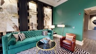 This stylish single-floor space makes a strong visual statement through its carefully curated interior design. The focal point of the room is a bold emerald green tufted velvet sofa, which complements the matching deep green accent wall, creating a cohesive and vivid color story. Contrasting the lush green tones, a rich cognac leather armchair adds warmth and texture to the seating arrangement.

A bold black-and-white geometric patterned area rug anchors the space, while a circular glass-top coffee table with a gold base introduces an element of glamour. The dark paneled feature wall serves as a dramatic backdrop for two large-scale framed art pieces with abstract, mixed-media compositions, framed and lit by dedicated picture lights that draw the eye upward.

Layered lighting, including tall floor lamps with white shades, ensures the space is well-illuminated while maintaining an intimate ambiance. Crown molding along the ceiling adds a refined architectural detail. The overall aesthetic blends Art Deco sensibilities with contemporary boldness, resulting in a sophisticated and visually dynamic environment.