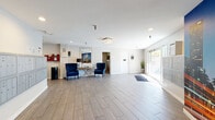 Located at 4355 Sepulveda Blvd in Los Angeles, this residential space offers a thoughtfully designed single-floor layout encompassing a living room and a generously sized bedroom. The main lobby serves as the heart of the common area, featuring wide-plank wood-look flooring that extends throughout, creating a warm and cohesive aesthetic. The lobby is bathed in natural light streaming through large glass doors and windows, enhancing the bright, airy atmosphere complemented by recessed lighting and a decorative flush-mount chandelier. A seating arrangement with elegant navy blue wingback chairs and a mirrored console table provides a welcoming focal point, while a striking floor-to-ceiling Los Angeles cityscape mural adds a bold, urban character to the space. A secondary lounge area features a tufted orange leather sofa and abstract wall art, offering a relaxed retreat alongside package and parcel locker stations for added convenience. The mailroom is lined with individual resident mailboxes and anchored by the same dramatic LA mural. Clean white walls, neutral gray tones, and consistent flooring throughout tie the spaces together, projecting a modern, well-maintained residential environment in the heart of Los Angeles.