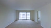 This single-floor space features a spacious, open living area characterized by its clean, minimalist design. The room is finished with neutral-toned wall-to-wall carpeting and crisp white walls, creating a bright and airy atmosphere. A prominent set of sliding glass doors dominates the far wall, fitted with vertical blinds that allow for adjustable natural light and provide a view of an outdoor area, likely a balcony or patio. The doors serve as the primary source of natural light, lending the room a soft, diffused brightness. The ceiling is smooth and standard height, contributing to the room's simple, understated aesthetic. White baseboards frame the perimeter of the room, adding a clean finish to the flooring transition. An open doorway on the right side of the living area leads to an additional room, suggesting a functional layout with clearly defined spaces. The overall ambiance is neutral and move-in ready, offering a blank canvas well-suited for a variety of interior design styles and furnishing arrangements.
