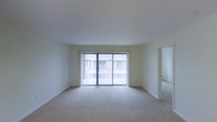 This single-floor space features a spacious, open living area characterized by its clean, minimalist design. The room is finished with neutral-toned wall-to-wall carpeting and crisp white walls, creating a bright and airy atmosphere. A prominent set of sliding glass doors dominates the far wall, fitted with vertical blinds that allow for adjustable natural light and provide a view of an outdoor area, likely a balcony or patio. The doors serve as the primary source of natural light, lending the room a soft, diffused brightness. The ceiling is smooth and standard height, contributing to the room's simple, understated aesthetic. White baseboards frame the perimeter of the room, adding a clean finish to the flooring transition. An open doorway on the right side of the living area leads to an additional room, suggesting a functional layout with clearly defined spaces. The overall ambiance is neutral and move-in ready, offering a blank canvas well-suited for a variety of interior design styles and furnishing arrangements.