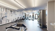 This modern fitness center offers a thoughtfully designed single-level layout that efficiently accommodates a comprehensive range of workout equipment. The space is defined by a striking, large-scale typographic mural spanning the main wall, featuring abstract lettering in muted grays and blues that lends an urban, energetic character to the interior.

Linear LED lighting runs along the ceiling, providing bright, even illumination throughout. On one end, floor-to-ceiling windows and glass doors flood the cardio zone with natural light and offer views to an outdoor area, creating an open, airy atmosphere. Multiple treadmills and upright cardio machines are positioned to take full advantage of this natural light.

Spin bikes are arranged on rubber mat flooring adjacent to the mural wall, while a dedicated free weights area includes a tiered dumbbell rack, adjustable bench, and a flat bench press. A dual-cable functional trainer occupies a secondary zone alongside a water fountain station and exit corridor. Stability balls and resistance accessories are neatly stored near the windowed entrance.

The durable rubber and speckled flooring throughout ensures safety and practicality, while the clean gray-and-white color palette maintains a polished, professional aesthetic suited for focused training.