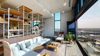 This elevated single-floor space combines refined design with an open, airy atmosphere, offering a seamless blend of collaborative and relaxation areas. Soaring ceilings and expansive floor-to-ceiling glass walls flood the interior with abundant natural light while framing sweeping panoramic views of the surrounding cityscape below.

The main lounge area is anchored by a striking open shelving unit crafted from warm wood and white metal framing, generously adorned with potted plants and decorative objects that bring a lively, biophilic energy to the space. Comfortable modular sofas with light upholstery and rattan detailing are arranged around low wooden coffee tables atop textured woven rugs, creating an inviting setting for casual gatherings or relaxed work sessions.

A dedicated meeting room features a distinctive perforated wood panel accent wall with a bold geometric cutout pattern, providing both visual interest and acoustic function. A large oval conference table surrounded by upholstered chairs sits beneath sculptural pendant lights, with city views visible through adjacent floor-to-ceiling windows. A curated art piece adds a creative accent.

The space extends onto a spacious outdoor terrace enclosed by a glass balustrade, where lounge seating and potted greenery take full advantage of the breathtaking open-air city views. Polished concrete flooring runs throughout, tying together the modern, urban aesthetic of the entire space.