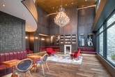 The Library at 695 Merrick Ave in Westbury, NY is a sophisticated and thoughtfully designed lounge space that blends classic elegance with contemporary style. The space is anchored by dramatic double-height ceilings adorned with an ornate gold coffered ceiling and a sweeping crystal chandelier that serves as a stunning focal point. Dark wood paneling lines the walls throughout, lending a warm, refined atmosphere reminiscent of a classic private library. Built-in bookshelves flank a marble fireplace, adding to the intellectual, club-like ambiance. The seating areas are furnished with rich crimson velvet and leather upholstered sofas, tufted benches, and uniquely designed accent chairs arranged atop an artfully patterned area rug. Brass accent tables and gold wall sconces add warm metallic touches throughout. A textured mosaic tile feature wall provides an eye-catching contrast to the wood paneling. Floor-to-ceiling steel-framed windows along one side of the space flood the interior with natural light while offering views of a lush outdoor courtyard, creating a seamless connection between the indoors and outdoors. Wide-plank hardwood flooring runs throughout, completing the space's polished yet inviting character.
