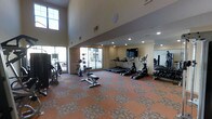 This fitness center is designed to support a comprehensive workout experience in a spacious, single-floor layout. The room's most striking architectural feature is its soaring double-height ceiling, which creates an open and airy atmosphere that enhances the overall sense of space. Large windows along one wall flood the interior with abundant natural light, offering views of the surrounding exterior and keeping the environment bright and inviting.

The facility is well-appointed with a variety of cardio equipment, including multiple treadmills and elliptical machines arranged in a neat row, all positioned facing a wall-mounted flat-screen television for entertainment during workouts. A dedicated free weights area includes a dumbbell rack stocked with a range of weights, complemented by a weight bench for strength training exercises. Additional cardio options, such as a stationary bike, round out the equipment selection.

The flooring features a durable, patterned carpet in warm terra cotta and neutral tones, adding texture and comfort underfoot. A mirrored wall on one side of the room amplifies the sense of space and allows users to monitor their form during exercise. Recessed ceiling lighting supplements the natural light, ensuring the space remains well-lit at all hours. Overall, the fitness center presents a clean, functional, and welcoming environment suited for a variety of fitness routines.