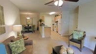 This single-floor residence features an open-concept layout that integrates the living, dining, and kitchen areas into one cohesive and functional space. The living area is carpeted and comfortably sized, anchored by a ceiling fan with a light fixture that provides both airflow and ambient lighting. A low coffee table and seating arrangement create a relaxed focal point, while natural green accents bring warmth to the room.

The dining area sits adjacent to the living space, defined by a pendant light overhead and a rounded table surrounded by green upholstered chairs, contributing a cohesive color palette throughout. A framed piece of wall art adds a decorative touch to this corner of the space.

The kitchen is separated from the living and dining areas by a peninsula counter with a dark countertop, offering additional prep space and a natural visual divider without closing off the open flow. Light wood cabinetry lines the kitchen walls, complemented by white appliances including a refrigerator and range. The kitchen floor transitions to hard surface flooring, contrasting with the carpet of the surrounding living areas. Overall, the space presents a functional, well-connected layout with a warm and inviting ambiance.