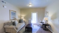 This single-story space centers around a thoughtfully designed living room that exudes a warm, inviting ambiance. The room features neutral beige walls that create a calm, versatile backdrop, complemented by carpeted flooring partially covered by a decorative area rug in soft blue and ivory tones. A flush-mount ceiling light provides consistent overhead illumination, while supplemental table lamps on either side of the seating arrangement add a warm, layered glow to the space.

The living room is furnished with a generously sized cream sofa accented by teal throw pillows, paired with a classic white wingback accent chair. A dark wood coffee table anchors the seating arrangement, offering both style and function. A framed landscape artwork on the left wall adds visual interest and a touch of color. On the right, a sleek side table with a glass-base lamp adds a contemporary accent.

A white-paneled exterior door at the far end of the room allows natural light to filter in through its window blind, enhancing the airy feel of the space. The overall design balances comfort and style, making the living area feel both functional and welcoming.
